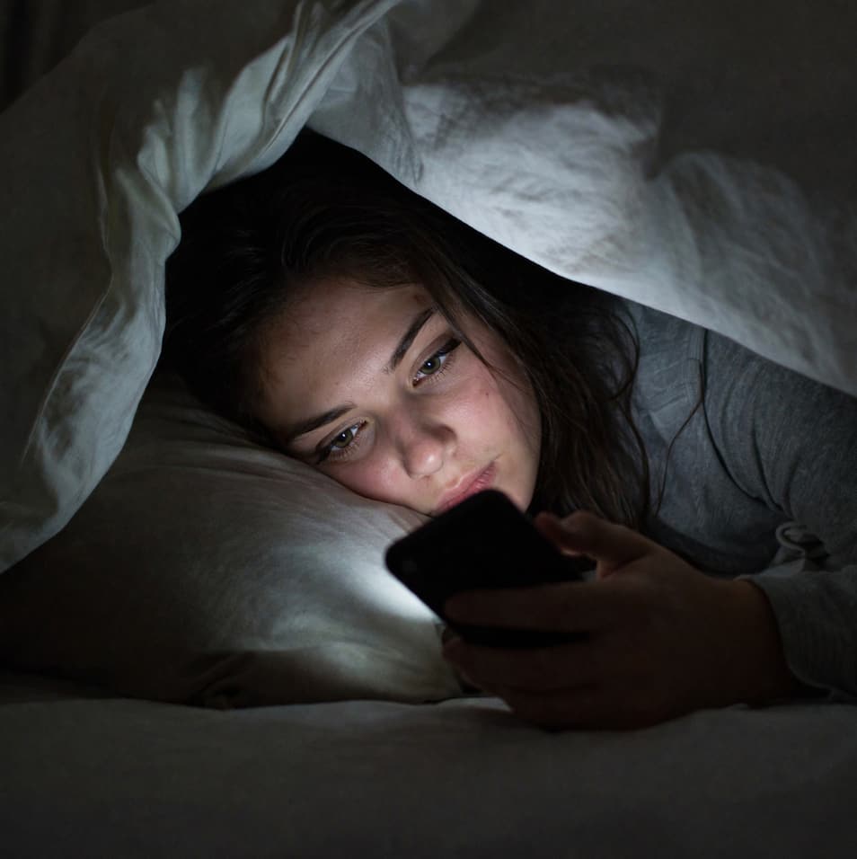 Kvinna ligger under täcket i ett mörkt sovrum och tittar diskret på sin mobil i skenet från skärmen
